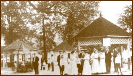 A sepia-toned photo of a group of people in vintage clothing near a gazebo in a park setting.