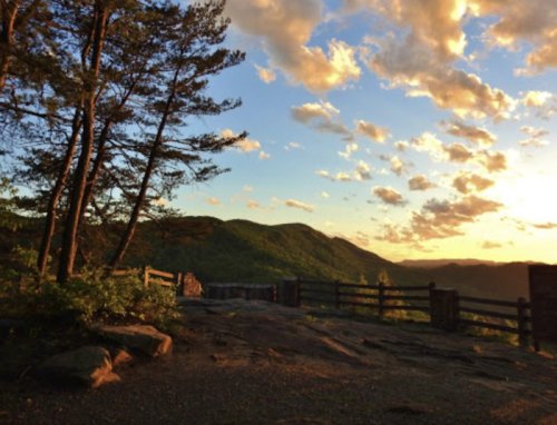 A scenic view of mountains at sunset, with trees and a wooden fence in the foreground. Soft clouds fill the sky.