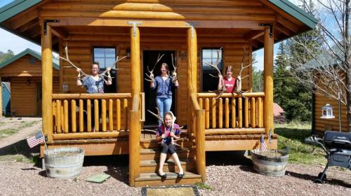 Four children stand on the porch of a log cabin, each holding sticks, smiling and posing for the photo.
