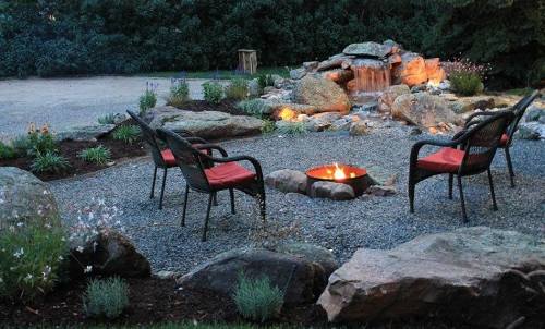 A cozy outdoor seating area with four chairs around a fire pit, surrounded by rocks and a small waterfall.