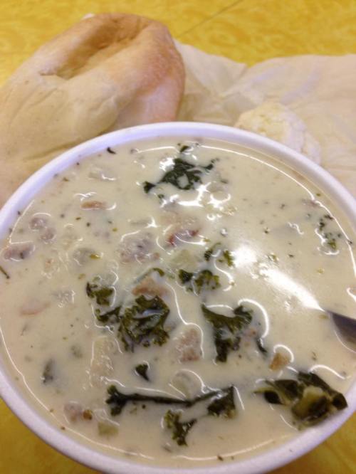 A bowl of creamy soup with greens and meat, accompanied by a roll and a biscuit on a yellow table.