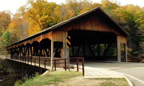 A wooden covered bridge spans a river, surrounded by autumn foliage in vibrant colors.