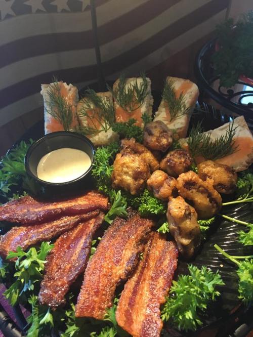 A platter featuring crispy bacon, chicken bites, and sushi rolls garnished with herbs and served with a dipping sauce.