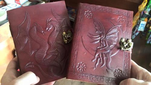 Two intricately designed leather journals with embossed dragon and fairy motifs, held in hands.