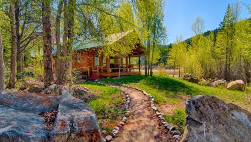 A scenic path leads to a wooden cabin surrounded by lush greenery and trees under a clear blue sky.