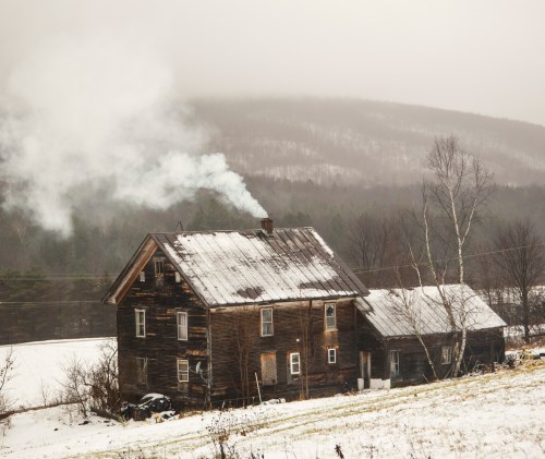 A rustic wooden house with smoke rising from the chimney, surrounded by snow and misty mountains in the background.