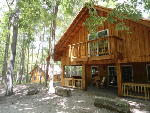 A wooden cabin surrounded by trees, featuring a balcony and a smaller cabin in the background.