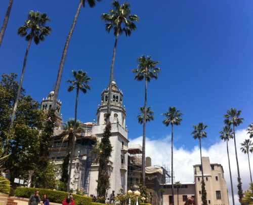 Historic building with towers, surrounded by tall palm trees under a clear blue sky. Construction scaffolding visible.