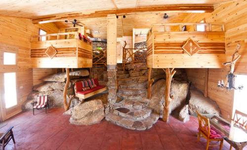 Interior of a rustic wooden cabin featuring two lofted beds, stone accents, and a cozy seating area.