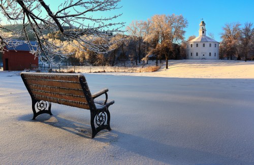 A frosty landscape featuring a wooden bench in the foreground and a white building with a clock tower in the background.