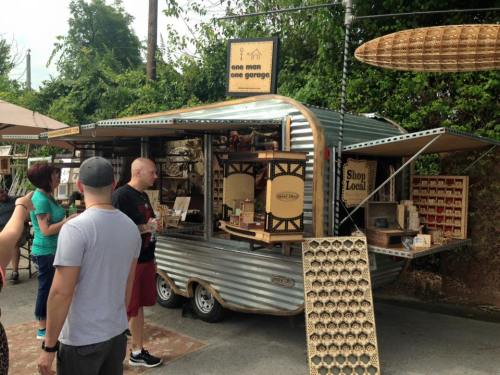 A vintage trailer shop with wooden crafts and decor, featuring customers browsing outside in a green, outdoor setting.