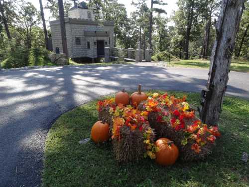 A cluster of pumpkins and autumn leaves on hay bales near a stone building surrounded by trees.