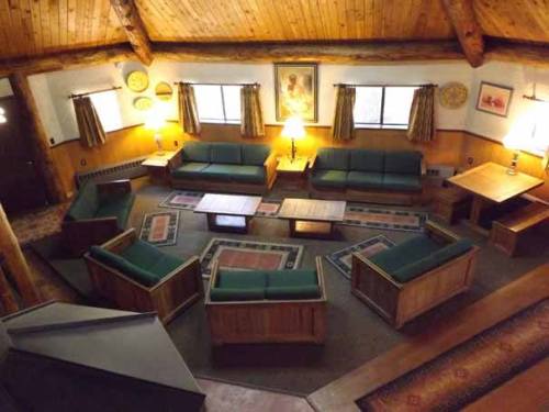 Cozy lodge interior with wooden beams, green sofas, and a central seating area surrounded by tables and rugs.