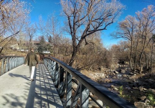 A person walks along a bridge beside a river, surrounded by bare trees and a clear blue sky.