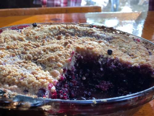 A close-up of a berry crumble pie with a golden crumb topping and a portion cut out, revealing the dark filling inside.