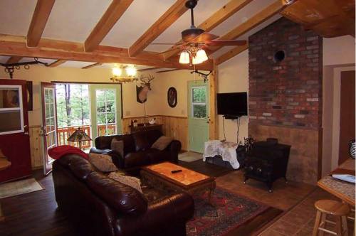 Cozy living room with wooden beams, a brick fireplace, leather sofa, and large windows overlooking a forested area.