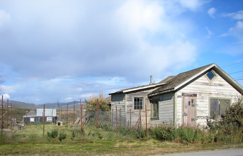 Abandoned house with weathered wood, surrounded by overgrown grass and a fence, under a cloudy blue sky.