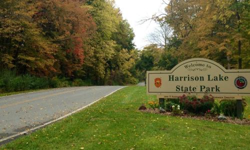 Welcome sign for Harrison Lake State Park, surrounded by trees with autumn foliage and a winding road.