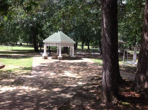 A white gazebo surrounded by trees in a park, with a paved path and grassy areas.