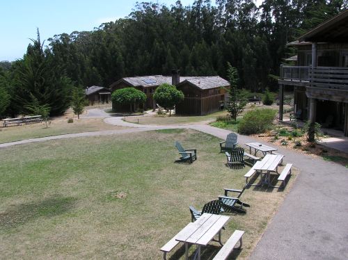 A serene outdoor area with wooden benches and chairs, surrounded by trees and rustic buildings.