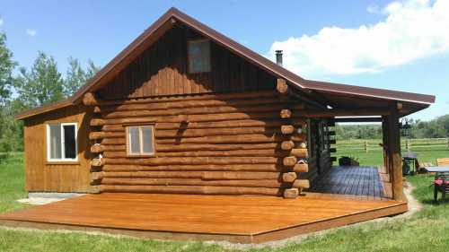 A rustic log cabin with a wooden deck, surrounded by green grass and trees under a blue sky.