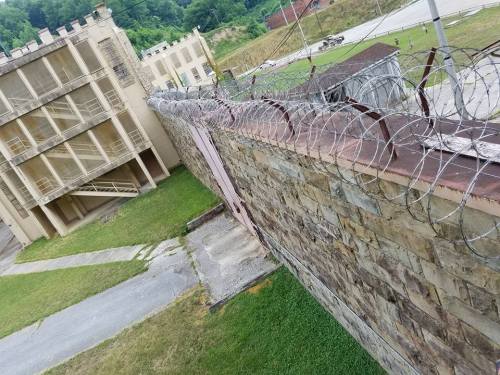 An aerial view of a stone wall topped with barbed wire, surrounding a dilapidated building and grassy area.