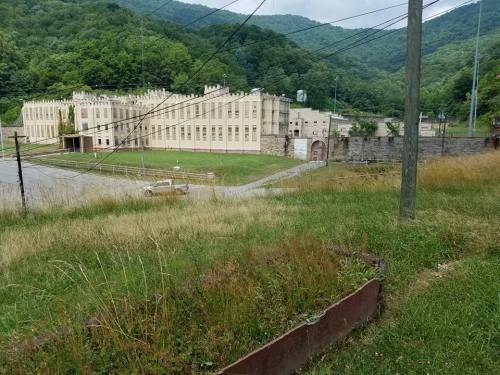 Abandoned building surrounded by overgrown grass and mountains in the background, with power lines in the foreground.