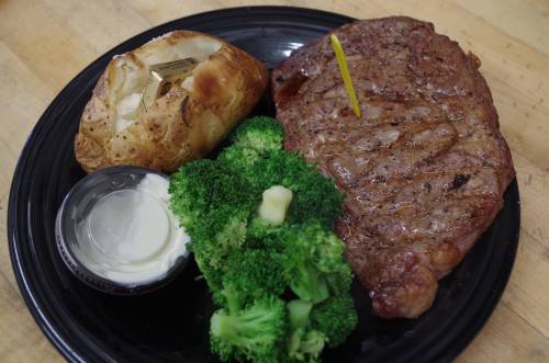 A plate with a grilled steak, baked potato, broccoli, and a small container of sour cream.