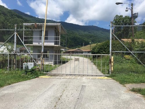 A gated entrance with a chain-link fence and a small building in the background, set against a mountainous landscape.