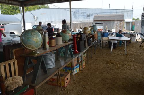 A market scene with tables displaying globes, vintage items, and people browsing in a rustic outdoor setting.