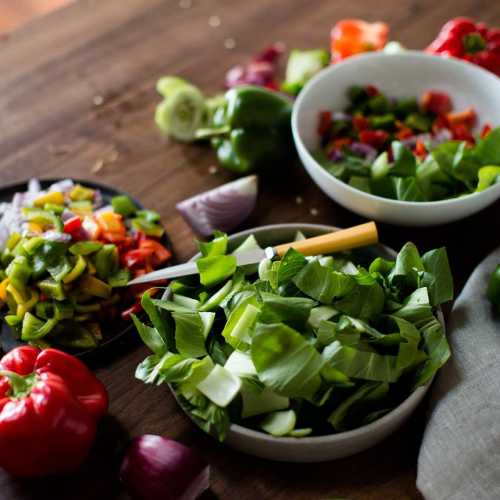 A colorful assortment of chopped vegetables, including bell peppers, onions, and leafy greens, on a wooden countertop.