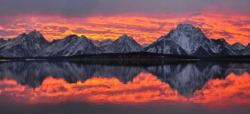 A stunning sunset over snow-capped mountains, reflecting in a calm lake with vibrant orange and purple hues in the sky.