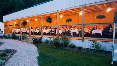 A dining area with guests seated at long tables, illuminated by warm lights, surrounded by greenery.