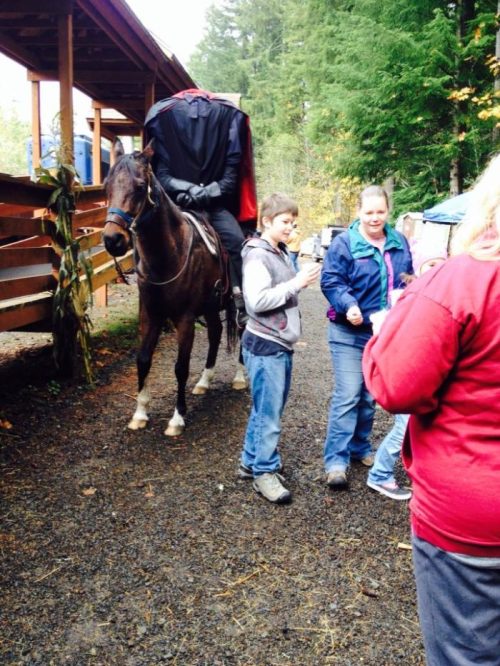 A group of people interacting near a horse, with a person in a cape sitting on the horse in a forested area.