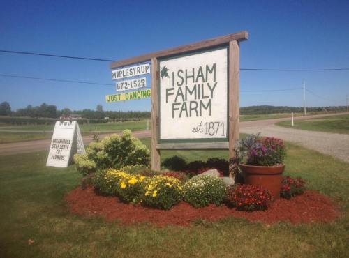 Sign for Sham Family Farm, established in 1871, with flowers and a road in the background. Nearby sign mentions maple syrup.