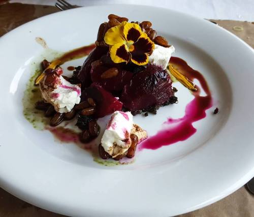 A gourmet dish featuring beets, goat cheese, seeds, and a flower garnish on a white plate with colorful sauces.