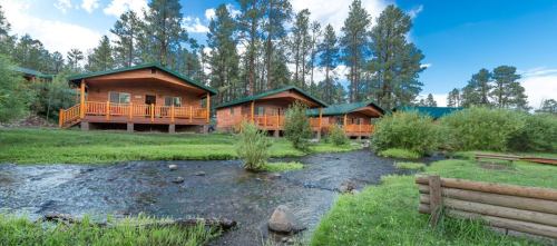 Three wooden cabins with green roofs beside a flowing stream, surrounded by trees and blue sky.