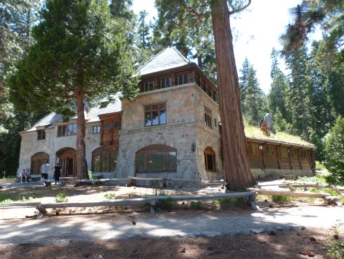 A large, rustic stone house surrounded by tall trees, with a grassy roof and people walking nearby.