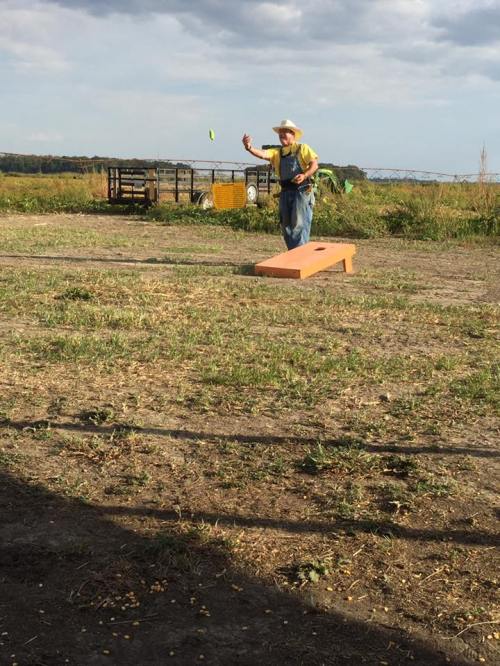 A person in a cowboy hat throws a bean bag towards a cornhole board in a grassy field under a cloudy sky.