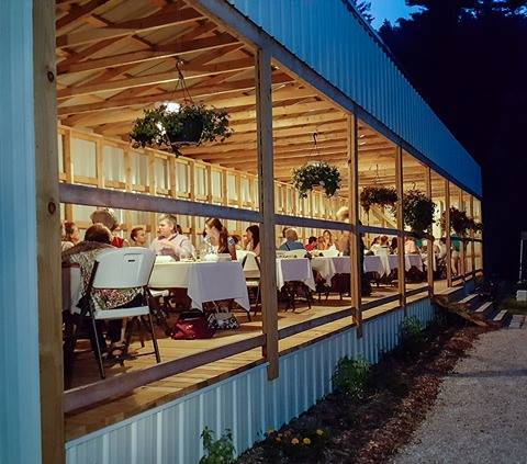 A gathering of people dining inside a well-lit, open-sided building at dusk, with tables set and plants hanging.