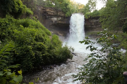A waterfall cascades down rocky cliffs, surrounded by lush greenery and flowing water below.