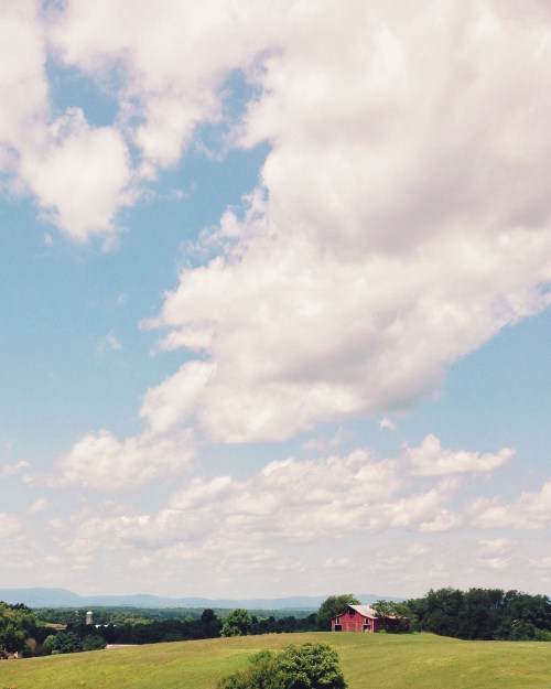 A scenic view of a green landscape with a red barn under a blue sky filled with fluffy white clouds.