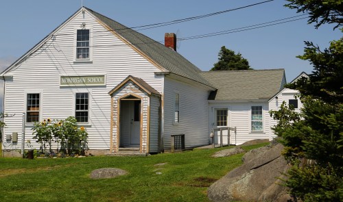 A white schoolhouse with a sloped roof, surrounded by greenery and rocks, under a clear blue sky.