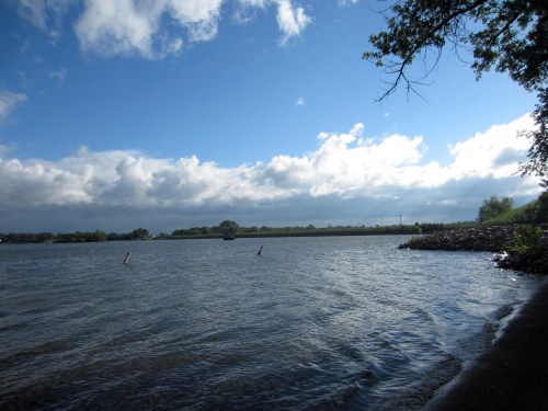 A serene river scene with gentle waves, under a partly cloudy sky and lush greenery along the shore.