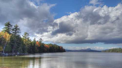 A serene lake surrounded by trees with autumn colors under a cloudy sky. Mountains are visible in the distance.