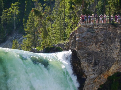 A waterfall cascades over a rocky ledge, with a group of people observing from a nearby viewpoint surrounded by trees.