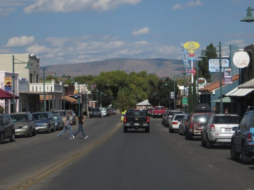 A small town street with shops, parked cars, and pedestrians crossing, set against a backdrop of mountains.
