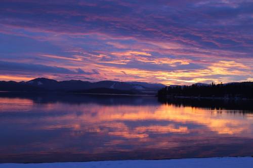 A serene lake at sunset, reflecting vibrant purple and orange hues in the sky and water, surrounded by mountains.