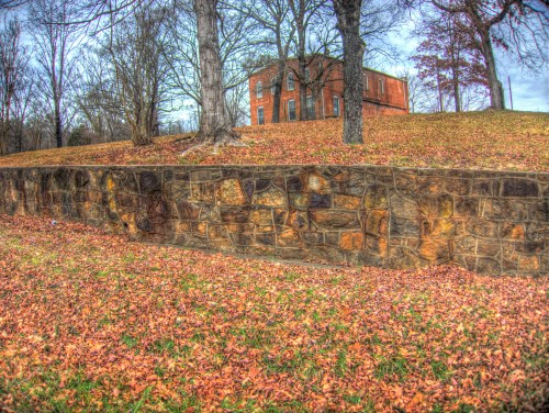 A stone wall in the foreground with a grassy area covered in autumn leaves and a brick building in the background.