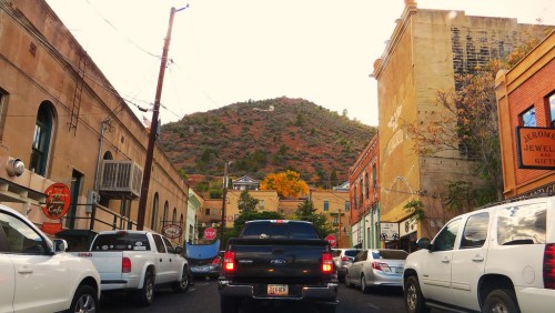 Traffic on a street lined with shops, with a mountain in the background and autumn foliage peeking through.